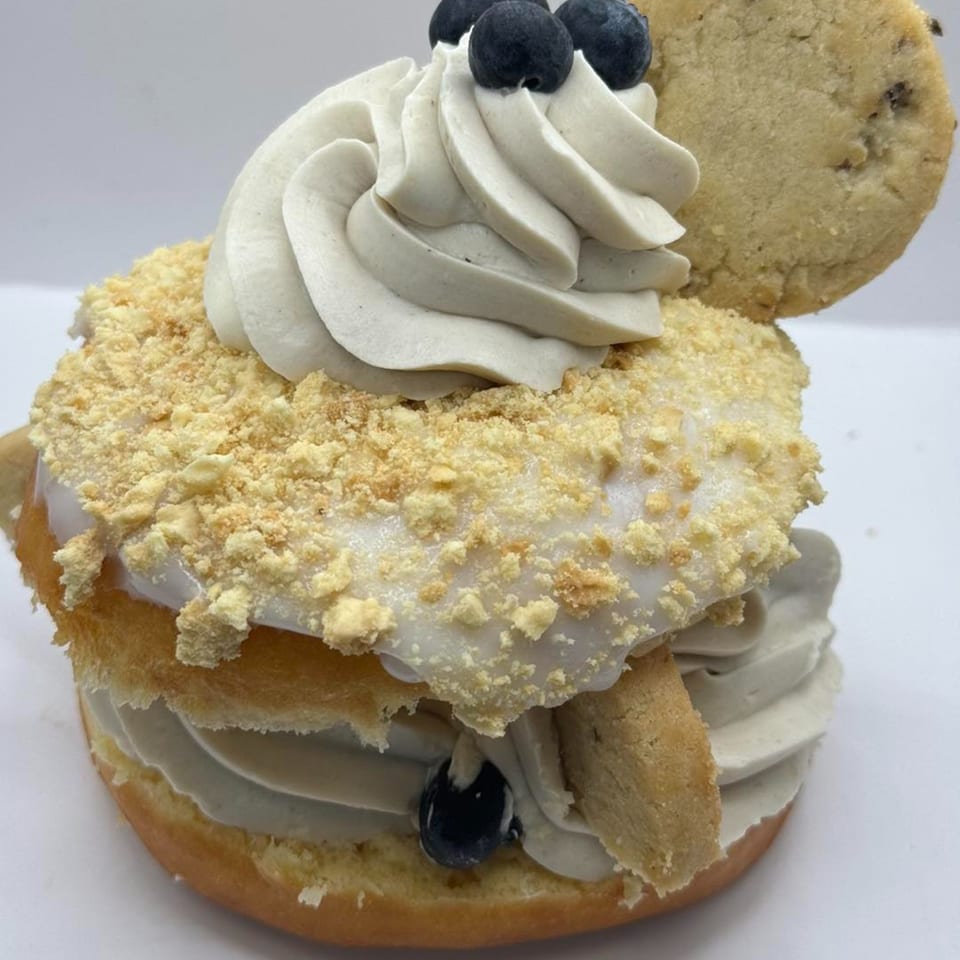 A Donut Factory donut with white icing, cookie crumbles, cream, blueberries, and a cookie.