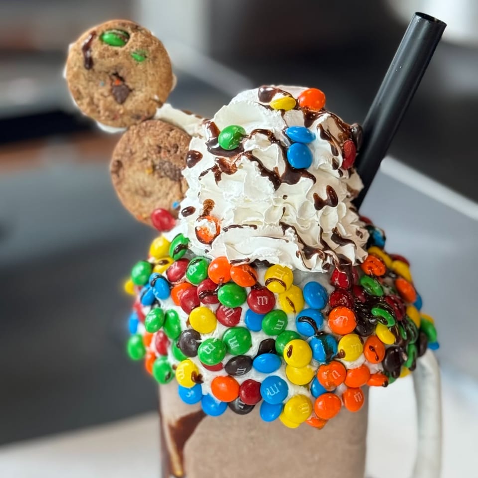 A Donut Factory milkshake overflowing with whipped cream, chocolate drizzle, M&M's, and two cookies.