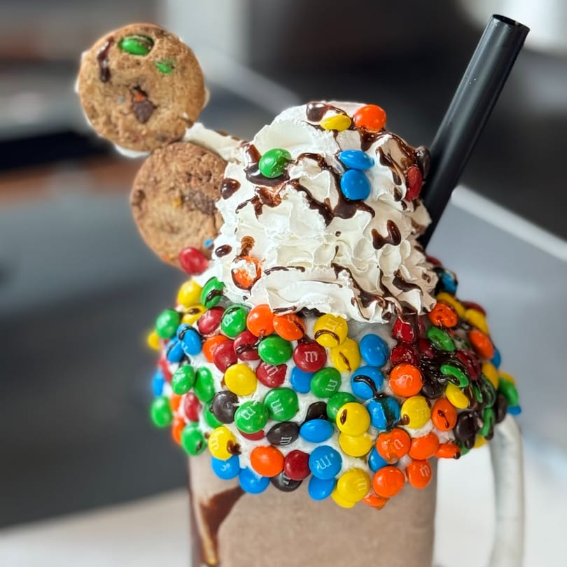A Donut Factory milkshake overflowing with whipped cream, chocolate drizzle, M&M's, and two cookies.