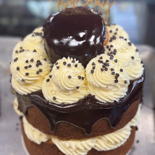 Chocolate layer cake with white frosting, chocolate drizzle, and mini chocolate chips, topped with a large glazed donut.