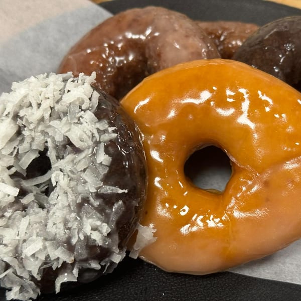 A chocolate donut with coconut flakes next to a glazed donut, with more donuts in the background.
