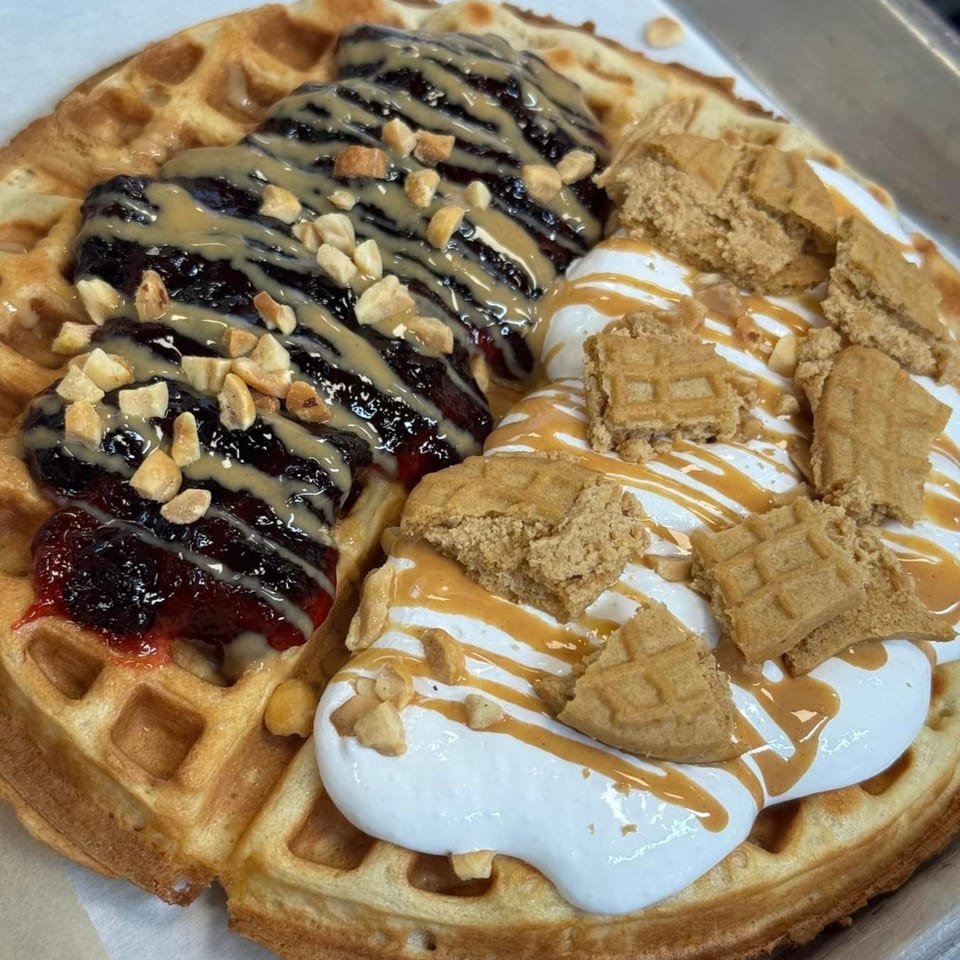A waffle topped with berry jam, peanut butter drizzle, whipped cream, and waffle pieces.