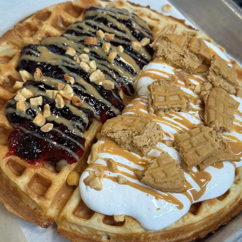 A waffle topped with berry jam, peanut butter drizzle, whipped cream, and waffle pieces.