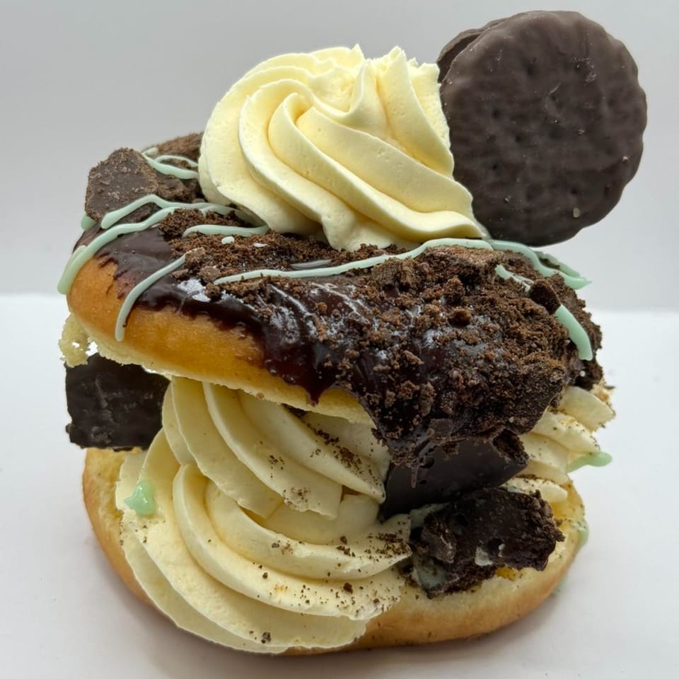 A stacked donut with chocolate glaze, mint drizzle, cookie crumbles, and whipped cream, topped with a chocolate cookie.