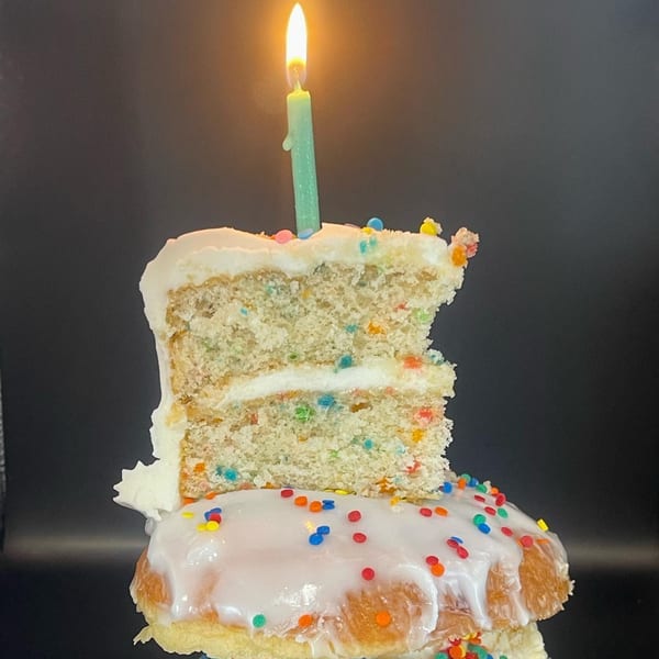 A slice of funfetti cake with a lit candle sits atop a glazed donut with sprinkles.